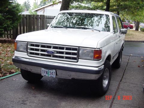 1987 Ford Bronco - White late-1980s Ford Bronco photographed head-on in a wet driveway, showing the bar-style grille with blue Ford oval, amber corner lamps a…