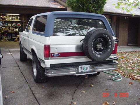 1987 Ford Bronco - Driver-side rear three-quarter view of a white full-size Ford Bronco with a blue hardtop, maroon tailgate stripe, chrome rear bumper and a…