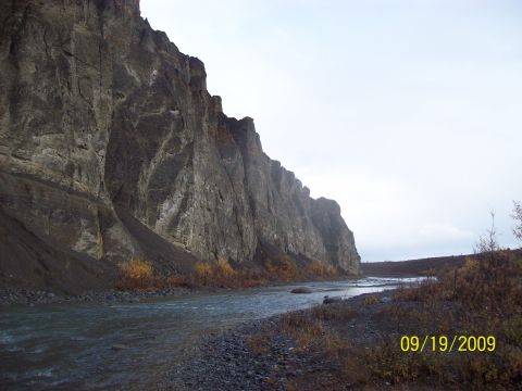 1986 Chevrolet Blazer - A wide river flows alongside a towering cliff face under an overcast sky.