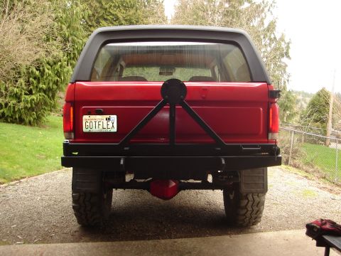 1989 Ford Bronco - Straight-on rear view of a red full-size Ford Bronco with a black removable hardtop, parked on a gravel and concrete driveway. It wears a f…