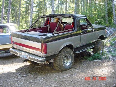 1989 Ford Bronco - A two-tone gray and black Ford Bronco with a roll cage is parked in a wooded area.