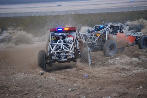 1982 Ford Bronco - Two off-road buggies, one with a red and blue light bar, kick up dust as they race on a desert track.