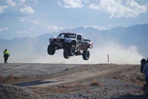 1982 Ford Bronco - A trophy truck soars through the air during a desert race, kicking up a large dust cloud.
