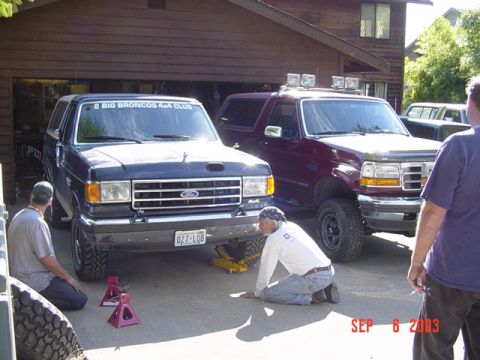 1989 Ford Bronco - Two Ford Broncos, one from the late 80s and a newer one, are in a driveway with men working on the older vehicle.