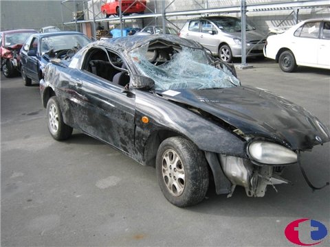 1993 Mazda MX-3 - A damaged black 1993 Mazda MX-3 coupe sits in a junkyard with other vehicles.