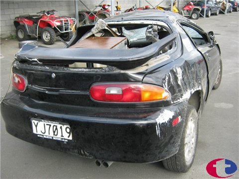 1993 Mazda MX-3 - This heavily damaged black coupe has a broken rear window and significant body damage, with debris visible inside.