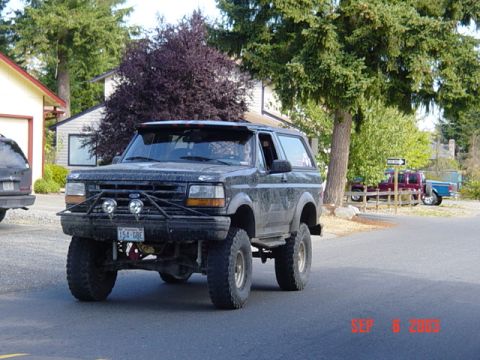 1989 Ford Bronco - A modified black Ford Bronco is seen on a street with houses and trees in the background.