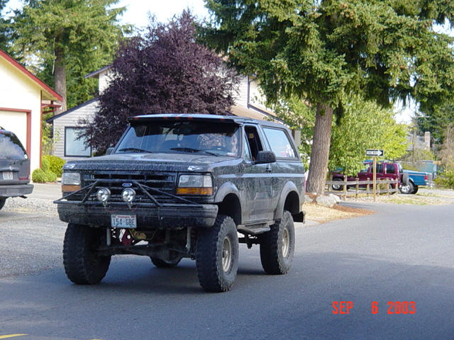 Mud-covered lifted Ford Bronco with tube bumper and driving lights on a residential street