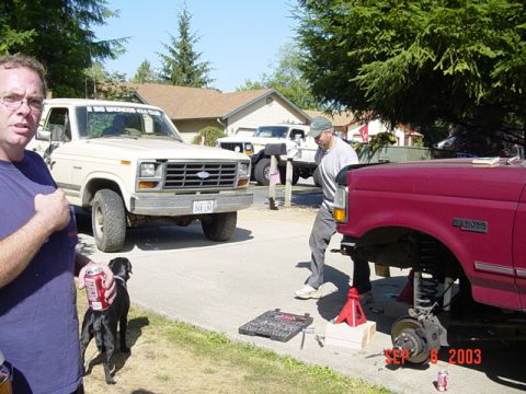 1989 Ford Bronco - Two men work on a red Ford truck in a driveway, with a cream-colored Bronco and another white SUV visible.