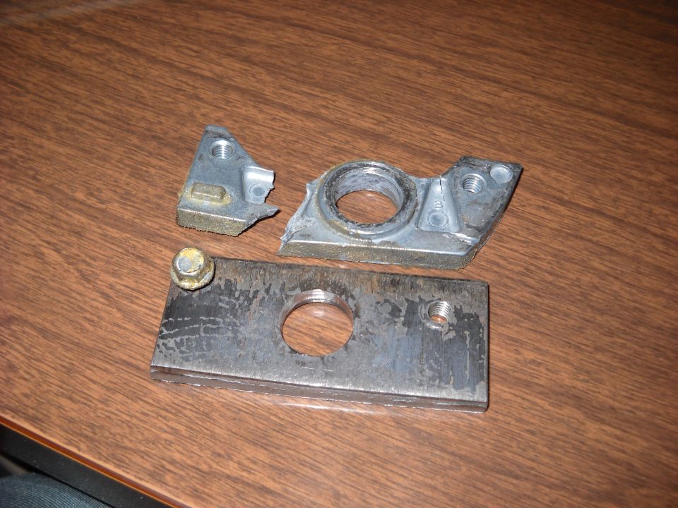 Broken metal bracket with a circular opening and a separate metal plate with a bolt. These parts are on a wooden surface