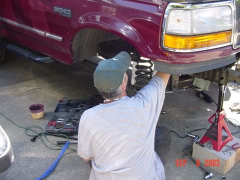 1989 Ford Bronco - A mechanic works on the front suspension of a maroon Ford Bronco, with tools and equipment laid out.