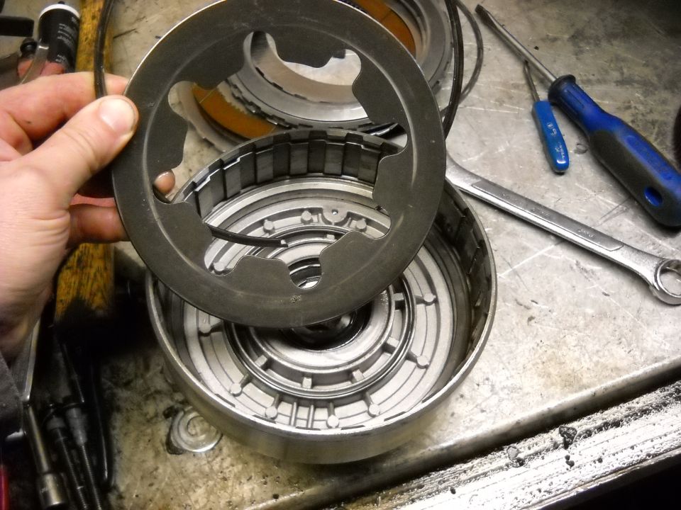Hand holding a metal clutch plate over a disassembled automatic transmission drum with tools on a workbench