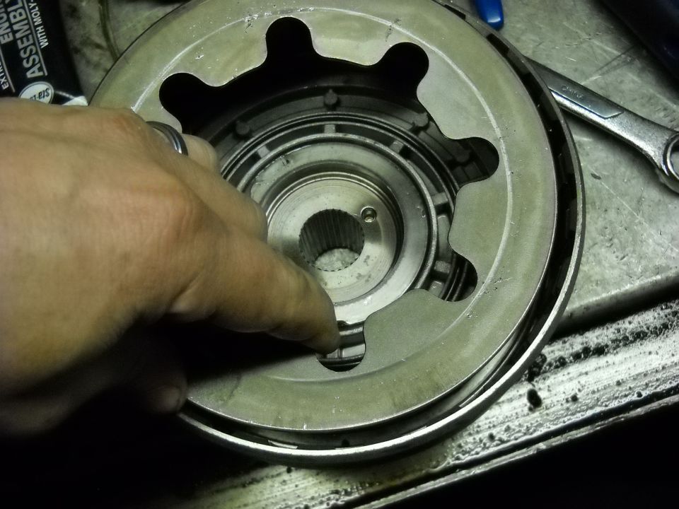 Hand holding a transmission clutch pack component. A wrench is visible in the background