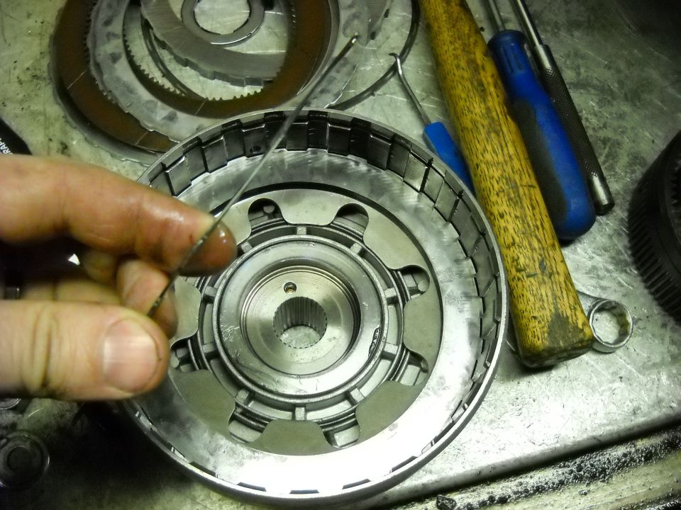 Hand holding a thin metal ring over a disassembled transmission clutch pack. Tools including a hammer and screwdrivers are visible
