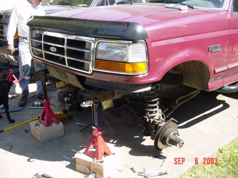 1989 Ford Bronco - Maroon Ford truck front end is disassembled for repair, with wheels off and supported by jack stands.