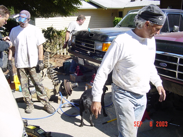 Black Ford truck on jack stands in a driveway with removed coil spring and rotor, people working