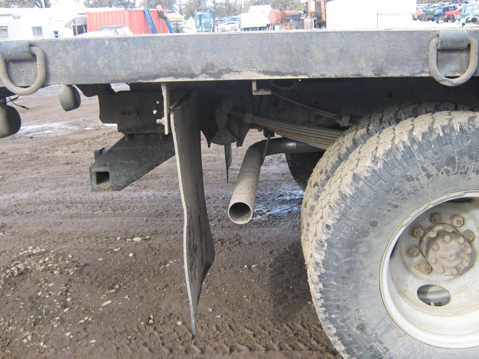 Truck chassis with dual rear wheels, mud flaps, and exhaust pipe. The vehicle is dirty and appears to be in a lot with other vehicles in the background