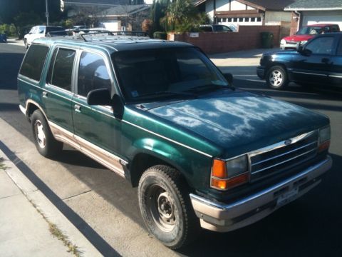 1993 Ford Taurus - A green SUV with a worn hood and tan trim is parked curbside, with other vehicles visible in the background.