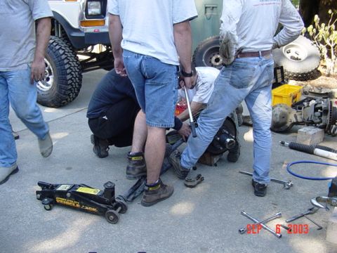 1989 Ford Bronco - Several people are gathered around a lifted truck, appearing to work on its front axle with tools and a jack nearby.