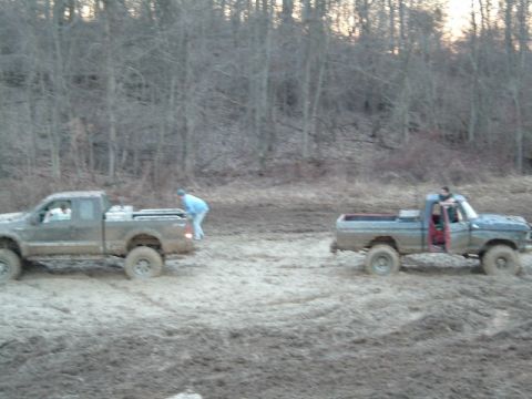 1993 Ford Bronco - Two pickup trucks are stuck in the mud with trees in the background.