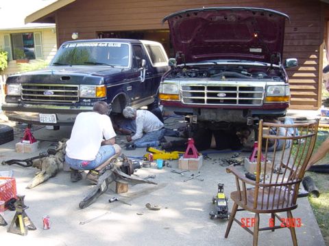 1989 Ford Bronco - Two Ford Broncos are undergoing maintenance outside a garage, with parts scattered on the ground.