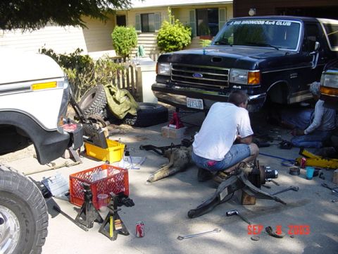 1989 Ford Bronco - Two people are working on a black Ford Bronco in a driveway, with a white truck also present. The scene is set up for repairs.