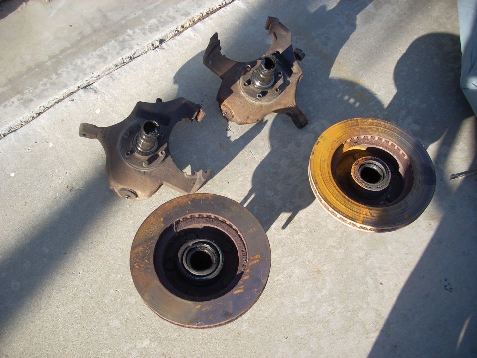 Two rusty brake rotors and two damaged steering knuckles on a concrete surface