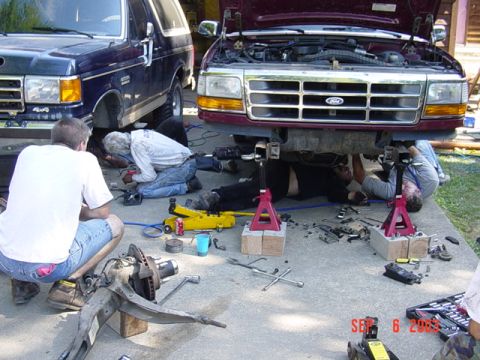 1989 Ford Bronco - Mechanics work on the front end of a maroon Ford Bronco, with another dark blue Bronco visible to the left.