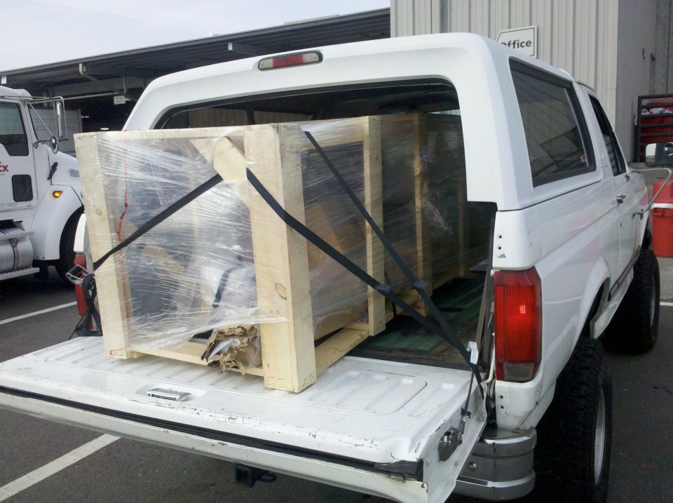 White lifted Ford Bronco with tailgate down hauling a strapped wooden crate at a freight dock