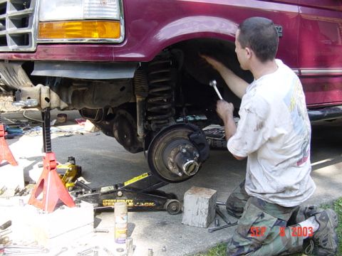 1989 Ford Bronco - A person works on the front suspension of a maroon Ford truck, which is supported by jack stands and a floor jack.
