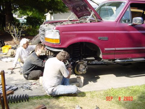 1989 Ford Bronco - Three individuals work on the front suspension of a maroon Ford Bronco, with the hood up.