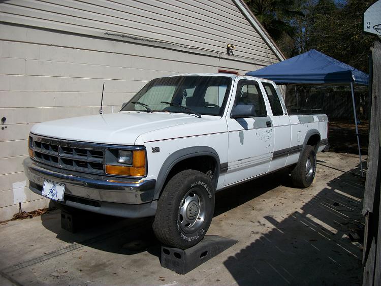 White Dodge Dakota extended cab pickup on wheel ramps, front three-quarter view by a block garage