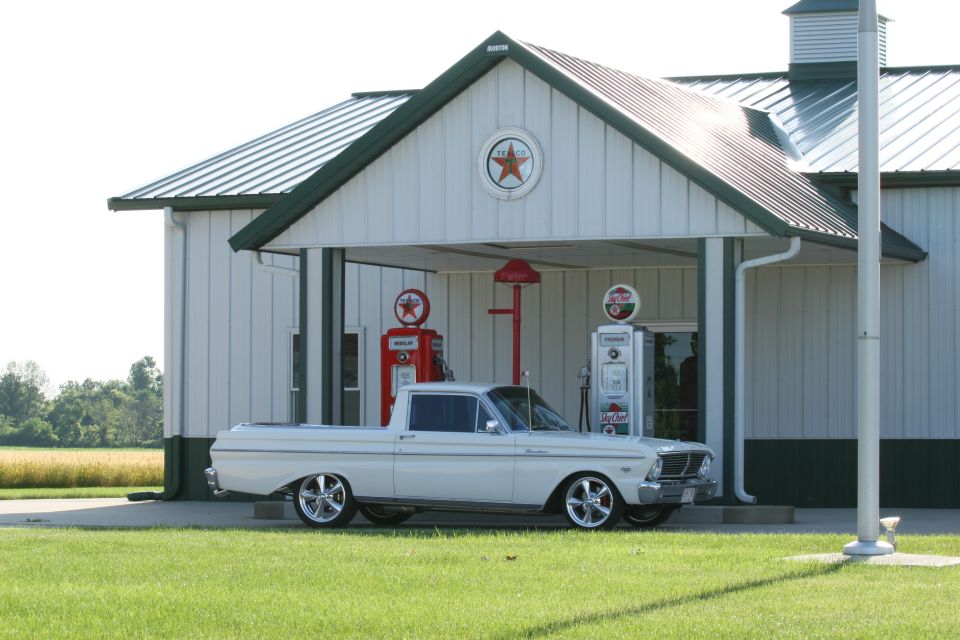 White vintage Ford Ranchero pickup parked before a Texaco-themed station with old gas pumps.