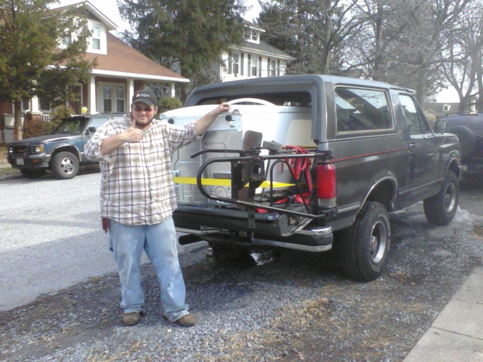 1988 Ford Bronco parked on gravel, with a man standing next to the open rear cargo area holding a large white item. Another vehicle is parked in the background