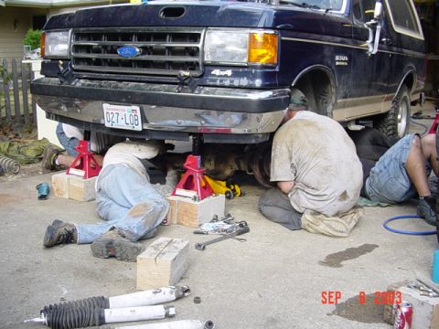 1989 Ford Bronco - Mechanics work on the front end of a dark blue 1989 Ford Bronco, supported by jack stands.