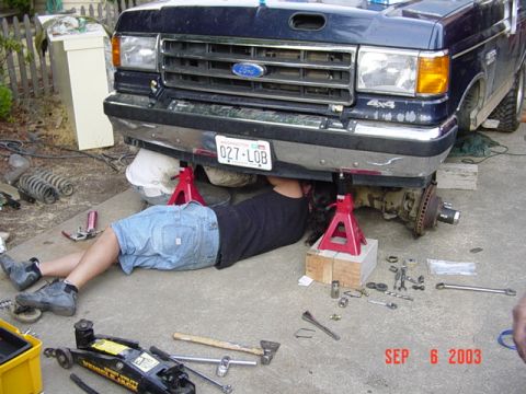 1989 Ford Bronco - A person works on the front suspension of a dark blue Ford F-Series truck, which is raised on jack stands.
