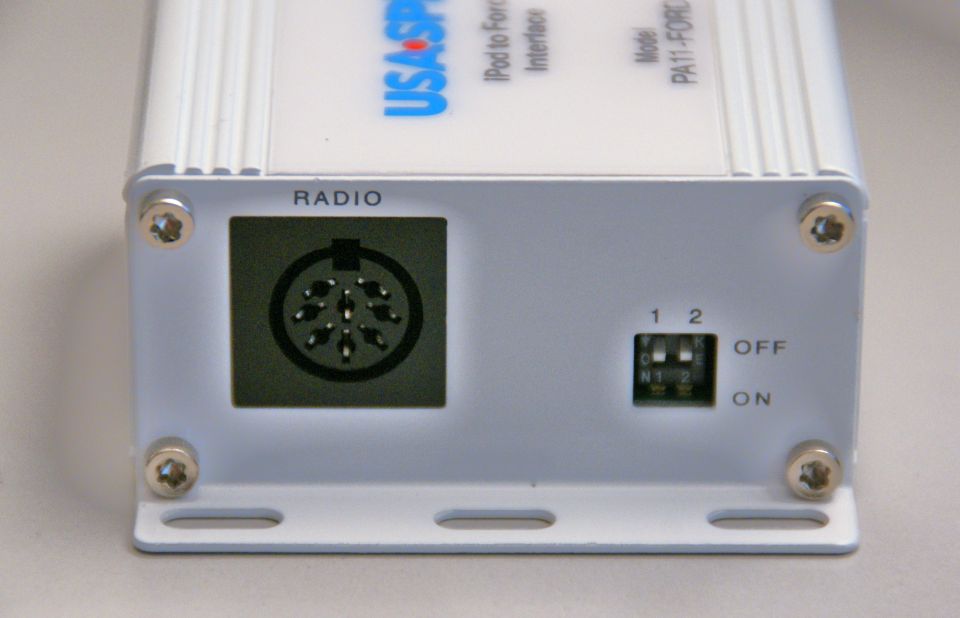 Close-up of a white electronic device with 'RADIO' port and DIP switches. Text reads 'USA SPEC iPod to Force Interface' and 'Model PA11-FORCE'