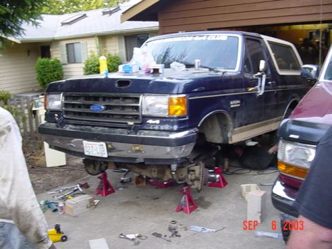 1989 Ford Bronco - A 1989 Ford Bronco is undergoing front-end work in a driveway, with tools and parts scattered around.