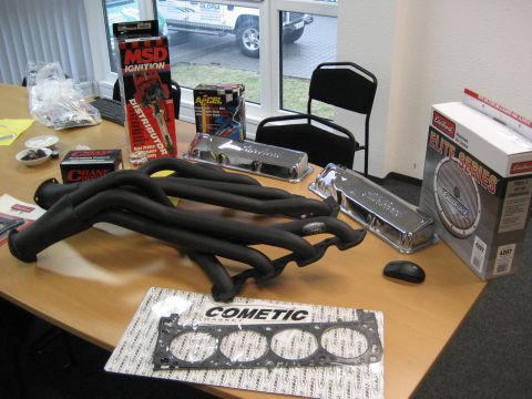 1973 Ford Ranchero - A haul of V8 speed parts spread across an office desk: black-coated long-tube headers, polished chrome valve covers, a Cometic head gasket…