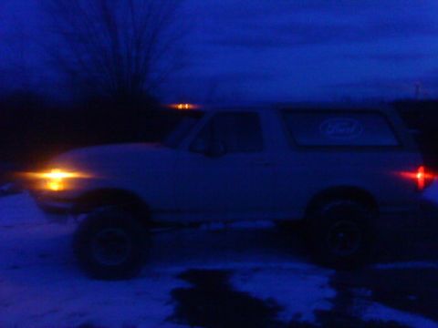 1995 Ford Bronco - A white Ford Bronco SUV is seen from the side at dusk, with its lights on.