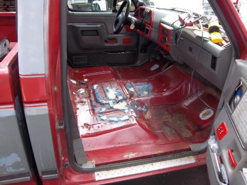 1989 Ford F-150 - The interior of a red pickup truck shows significant wear and tear on the floor, with carpet removed.