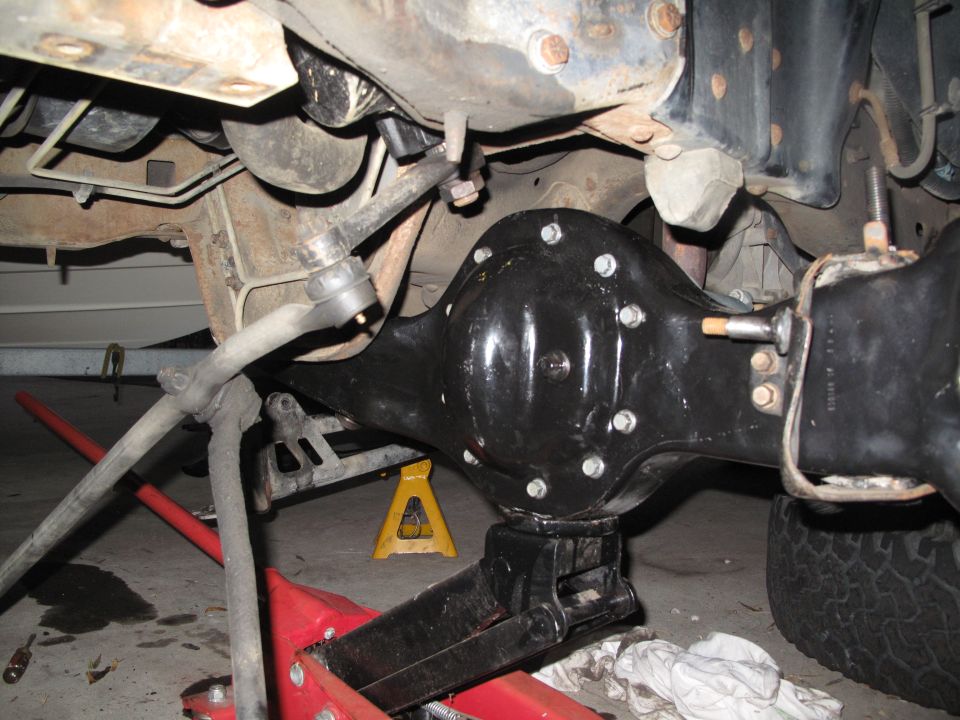 Close-up of a vehicle's rear axle assembly, supported by a jack and jack stands. Steering linkage is visible