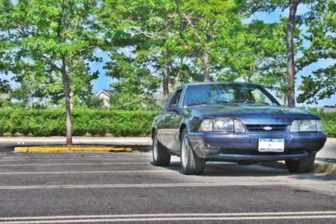 1993 Ford Mustang - A dark blue coupe is parked in a lot, with trees and bushes in the background.