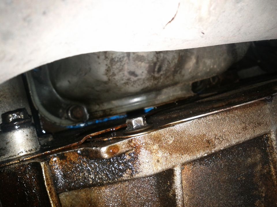 Close-up of greasy engine oil pan and transmission housing with bolts and blue sealant visible