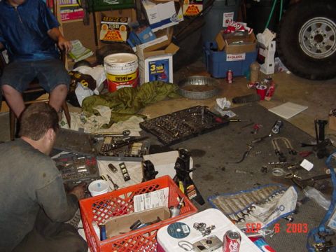 1989 Ford Bronco - A cluttered garage scene shows two individuals engaged in vehicle maintenance, with tools and parts scattered around.