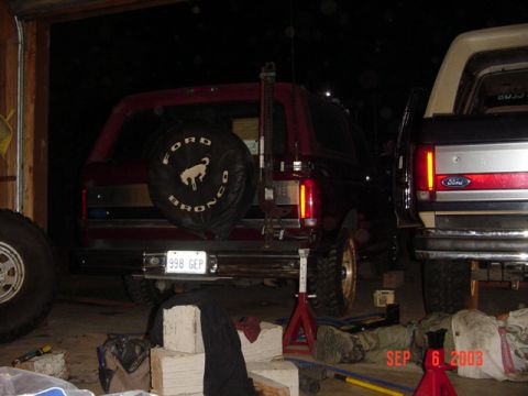 1989 Ford Bronco - A red 1989 Ford Bronco is parked next to another vehicle, showing its rear end and spare tire cover.