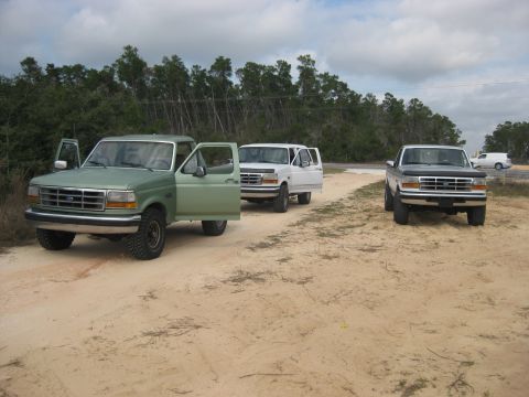 1994 Ford Bronco - Three Ford trucks, including two F-150s and a van, are parked on a dirt road with trees in the background.