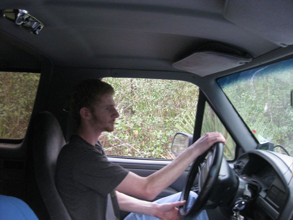 Interior view of a man driving a vehicle, with trees and brush visible through the windows