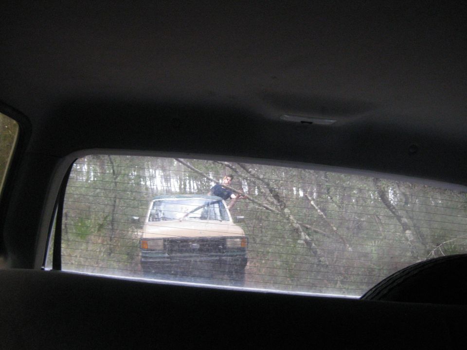 Tan SUV with a person on its roof, seen through a dirty rear window