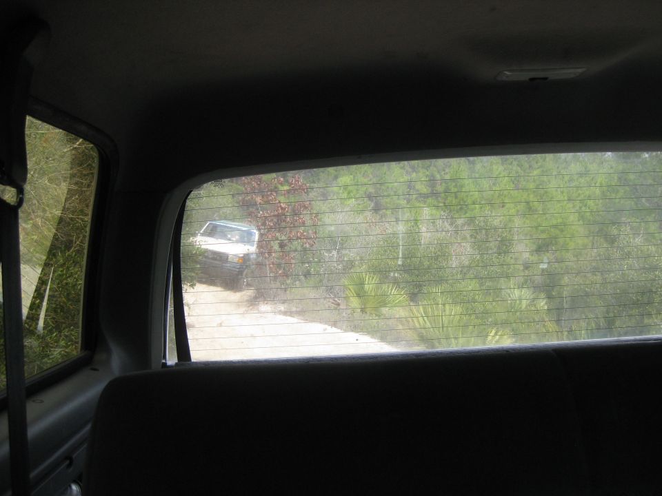 White SUV driving on a dirt road through trees, seen from inside another vehicle